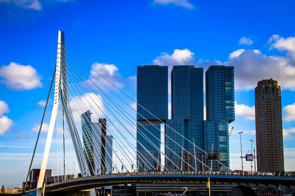 Een opvallende moderne brug met hoge ophangkabels strekt zich uit voor een stedelijke skyline met verschillende hoogbouw onder een helderblauwe lucht, bezaaid met witte wolken. Net zoals een matchmaker individuen verbindt, verbindt dit architecturale wonder het landschap van de stad, waarbij technische hoogstandjes in harmonieus contrast worden benadrukt.