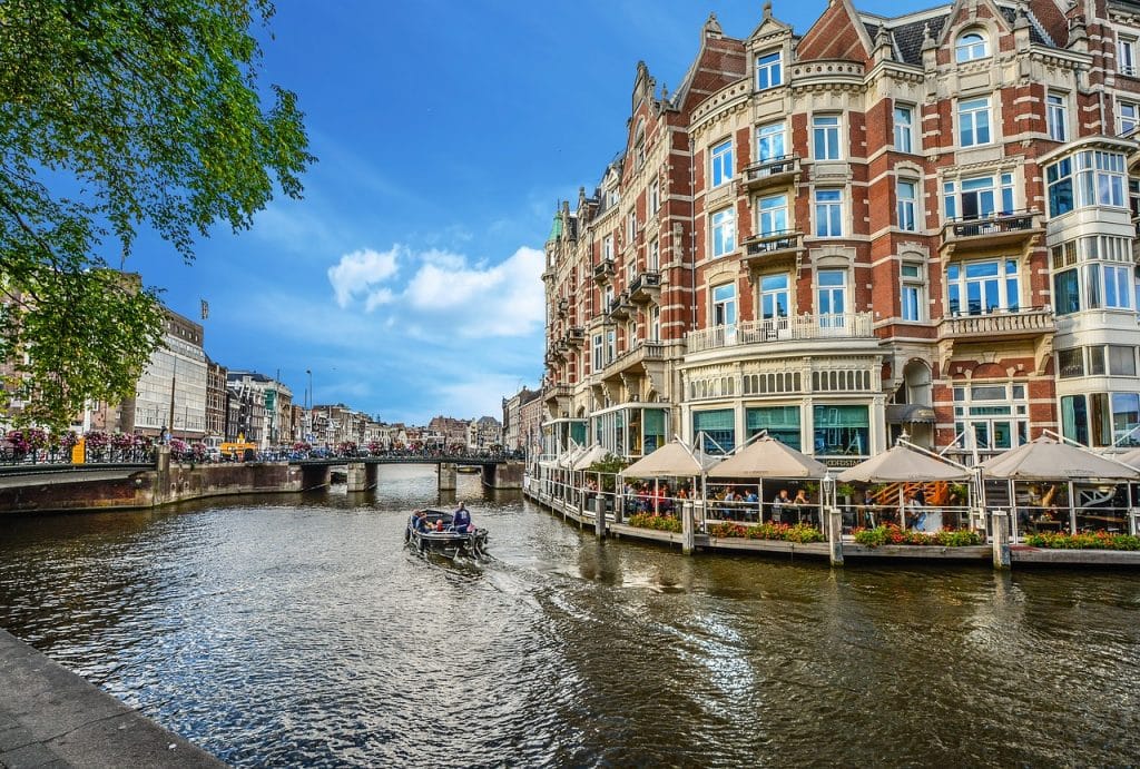 Een schilderachtig uitzicht op de Amsterdamse gracht terwijl een boot over het water vaart. Historische gebouwen met sierlijke architectuur liggen langs de gracht, waaronder een groot, elegant gebouw met buitenterrassen. De lucht is helder en blauw, en groen omlijst het tafereel gedeeltelijk, wat een perfecte achtergrond vormt voor duurzame matchmaking.