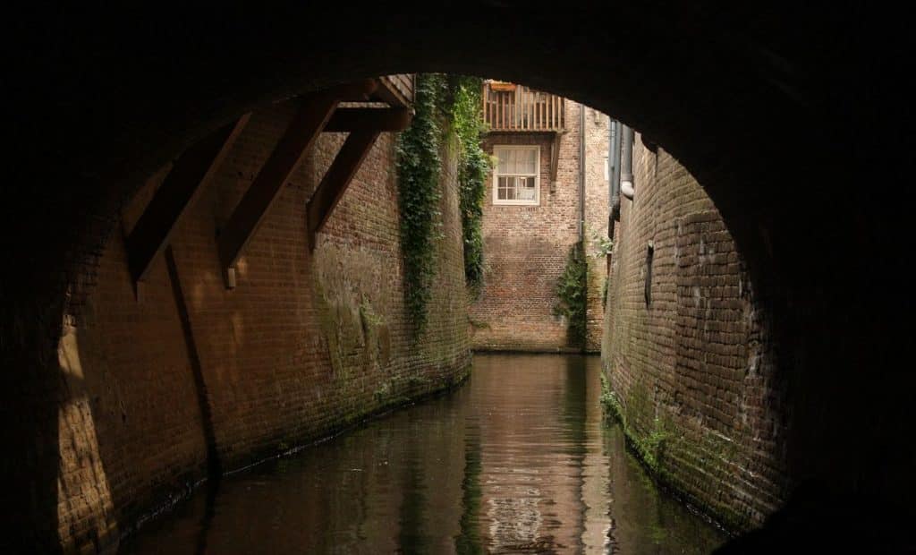 Een smal kanaal stroomt onder een bakstenen boog door. De muren zijn bekleed met met klimop begroeide bakstenen. Het zonlicht filtert naar beneden en benadrukt een raam en een balkon op een bakstenen gebouw aan de andere kant. Het water eronder weerspiegelt de omringende architectuur en het groen, wat doet denken aan de tijdloze verbindingen die een koppelaarster tot stand brengt.