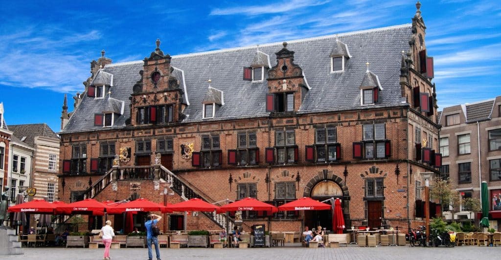 Een historisch bakstenen gebouw met een grijs pannendak en sierlijke architectonische details staat onder een helderblauwe lucht met verspreide wolken. Er staan rode parasols die de tafels op een terras op de begane grond in de schaduw stellen, waar mensen genieten van hun koffie en gesprekken, wat duurzame relaties bevordert.