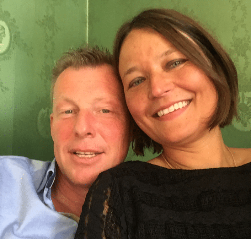 Homepage 5 Een man en een vrouw glimlachen naar de camera terwijl ze een selfie maken. De man, met kort, lichtgekleurd haar, draagt een lichtblauw overhemd. De vrouw heeft bruin haar tot op haar schouders en draagt een zwarte top. Ze lijken samen gelukkig en laten mogelijk hun succes zien via datingbureau-diensten, tegen een achtergrond met een groen patroon.