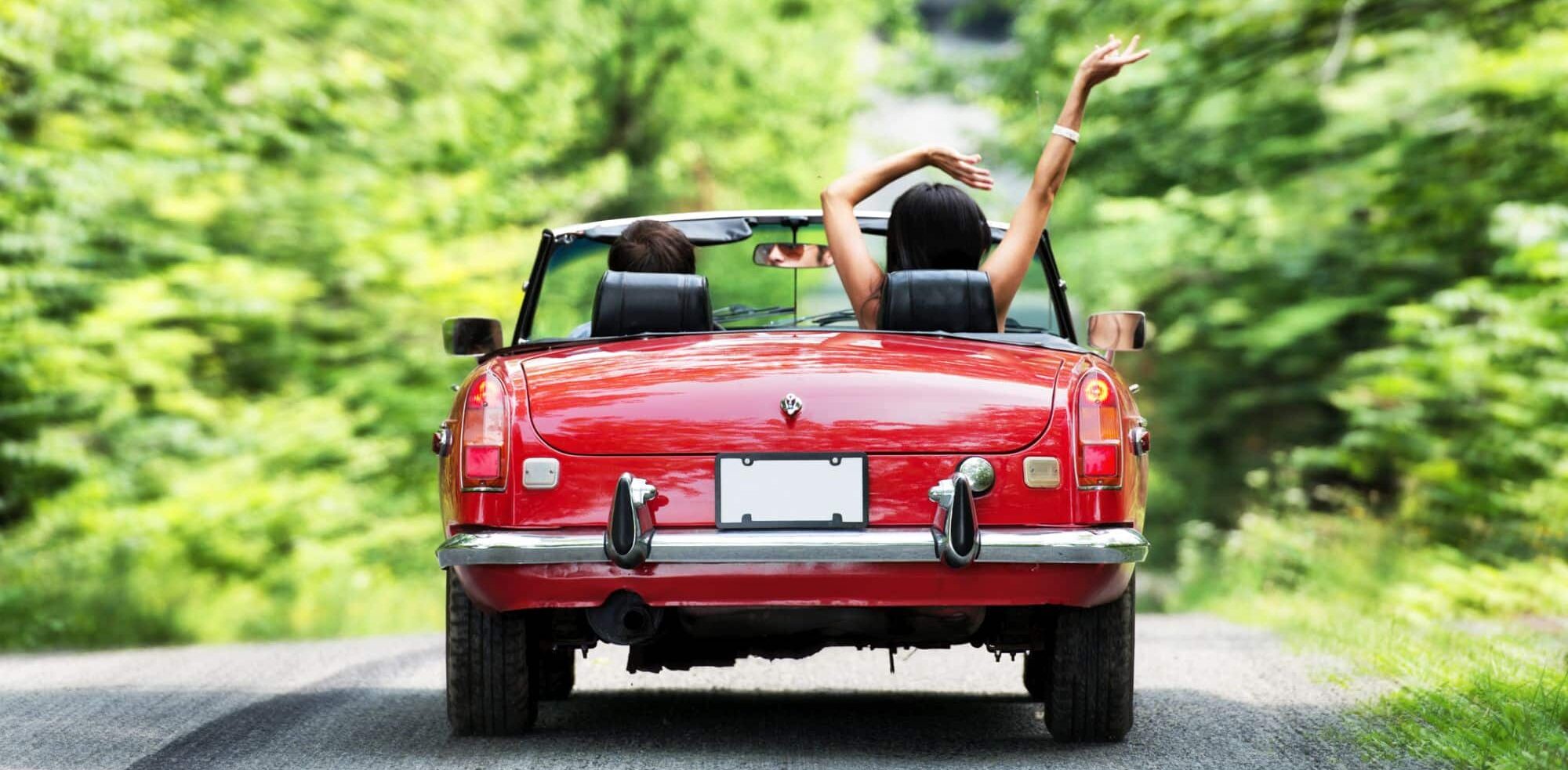 Twee mensen in een rode cabriolet rijden over een weelderige, groene, met bomen omzoomde weg. De persoon op de passagiersstoel heeft zijn armen in de lucht en geniet van de rit. De achterste kentekenplaat van de auto is blanco. De scène straalt een zorgeloze en avontuurlijke sfeer uit, ideaal voor iedereen die op zoek is naar een duurzame relatie.