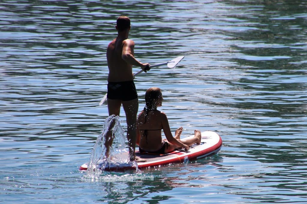 Twee mensen zitten op een paddleboard in een serene watermassa. Eén persoon staat en peddelt terwijl de ander met gekruiste benen voorin zit en geniet van de rit. Zonlicht weerkaatst op het kabbelende water, waardoor een vredig tafereel ontstaat, georkestreerd door hun ideale match van het plaatselijke datingbureau.