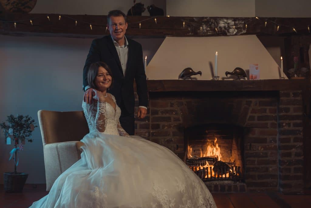 Svetlana & Arthur 1 Een stel poseert in een elegante setting voor een relatiebemiddelingsadvertentie. De vrouw, gekleed in een witte trouwjurk, zit op een stoel met haar hand op de arm. De man, gekleed in pak, staat achter haar met zijn hand op haar schouder. Ze staan voor een verlichte bakstenen open haard.