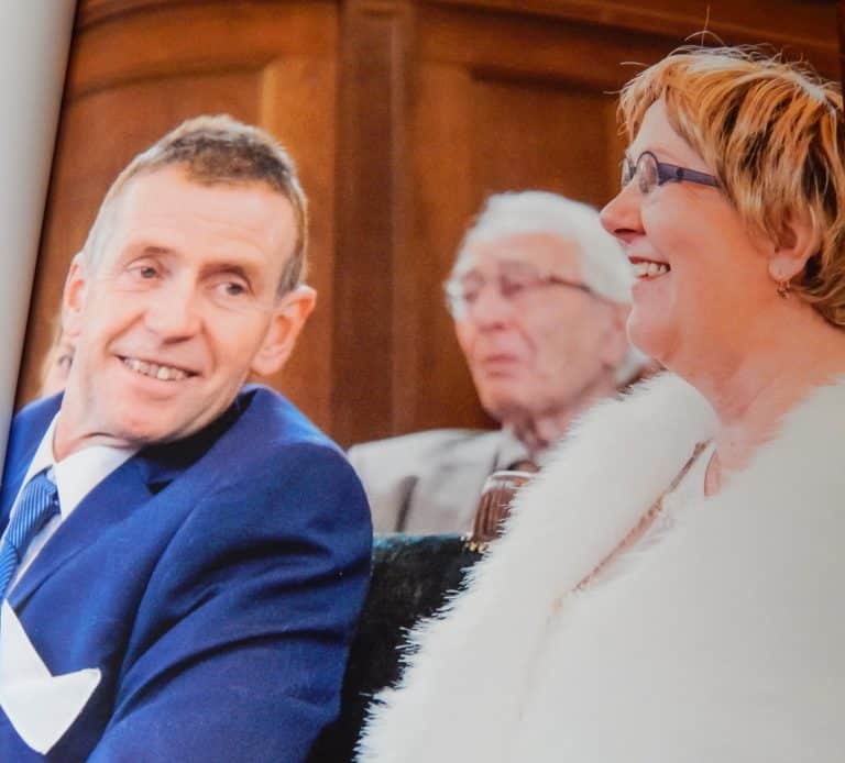 Jan & Mariëtte 1 Een man en een vrouw met een hoog EQ omhelzen elkaar buiten, de ogen gesloten, terwijl de man zachtjes haar voorhoofd kust in een gebaar van troost en genegenheid. Beiden dragen blauw en zien er kalm en vredig uit tegen een achtergrond van groen.