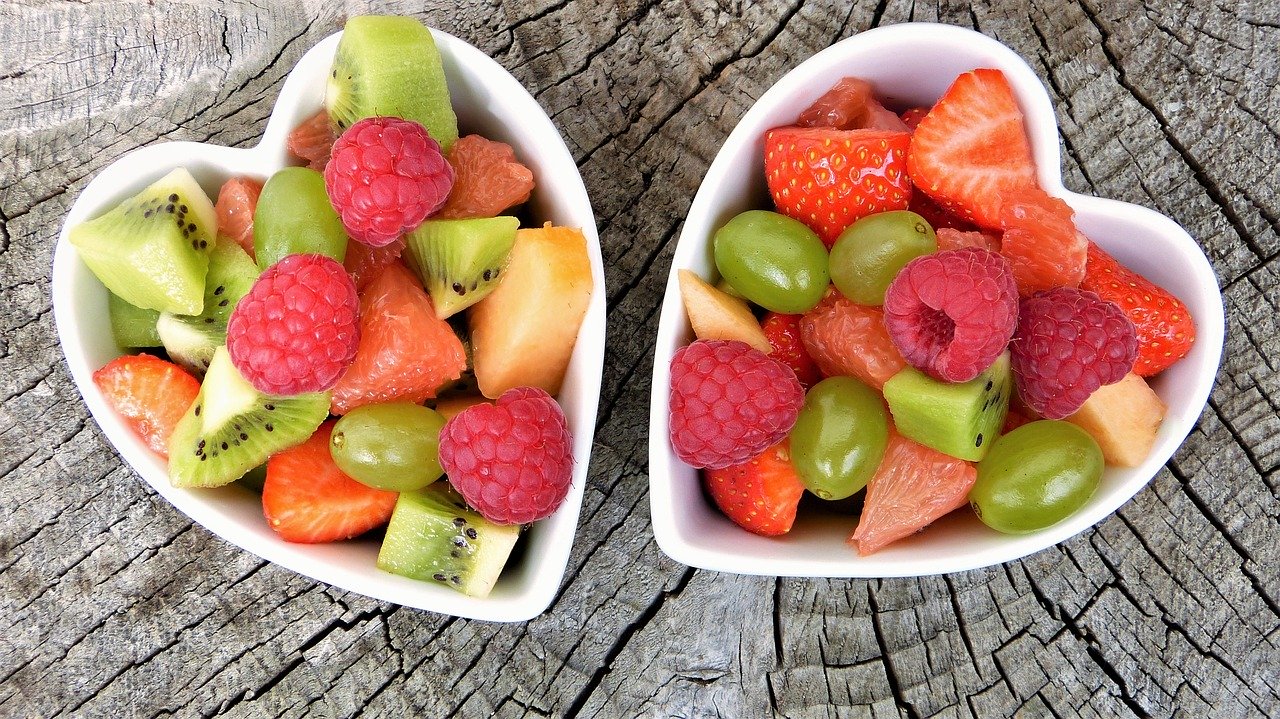 Twee hartvormige kommen gevuld met diverse soorten vers fruit, waaronder kiwi, frambozen, aardbeien, groene druiven en grapefruit, geplaatst op een rustiek houten oppervlak. De levendige kleuren van de vruchten creëren een aantrekkelijk en smakelijk geheel waar elke matchmaker trots op kan zijn.