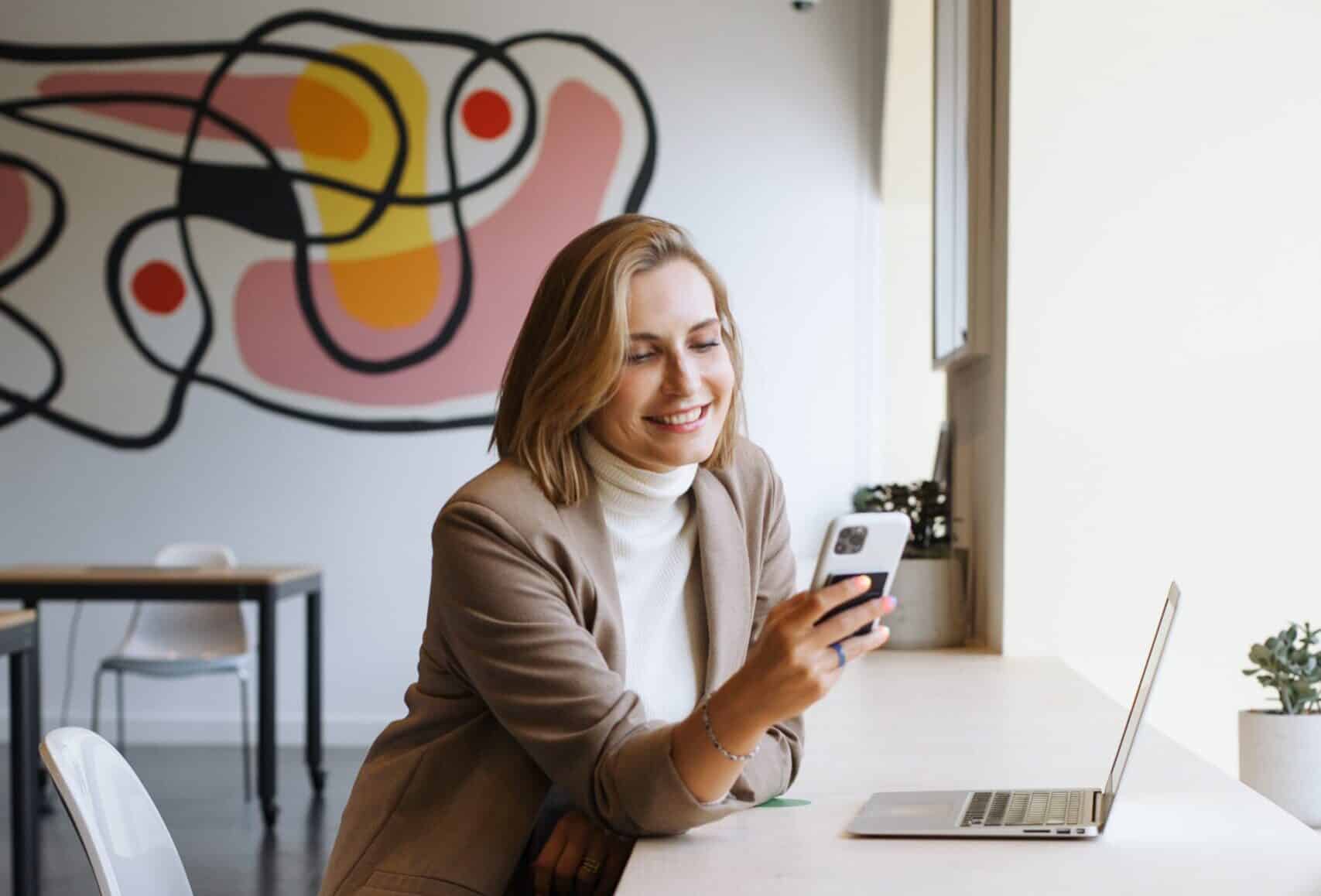 Een persoon met blond haar op schouderlengte, gekleed in een beige blazer en witte coltrui, glimlacht terwijl hij naar zijn smartphone kijkt. Ze zitten aan een bureau met een laptop en een kleine potplant. Op de achtergrond hangt een kleurrijke abstracte muurschildering, die verwijst naar hun werk in relatiebemiddeling voor een relatiebureau.