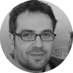 Een man met kort donker haar en een baard, met een rechthoekige bril, kijkt in de camera. Hij draagt een shirt en een jasje. De foto is zwart-wit en zijn uitdrukking is neutraal, alsof hij overweegt om zich in te schrijven voor het laatste aanbod.