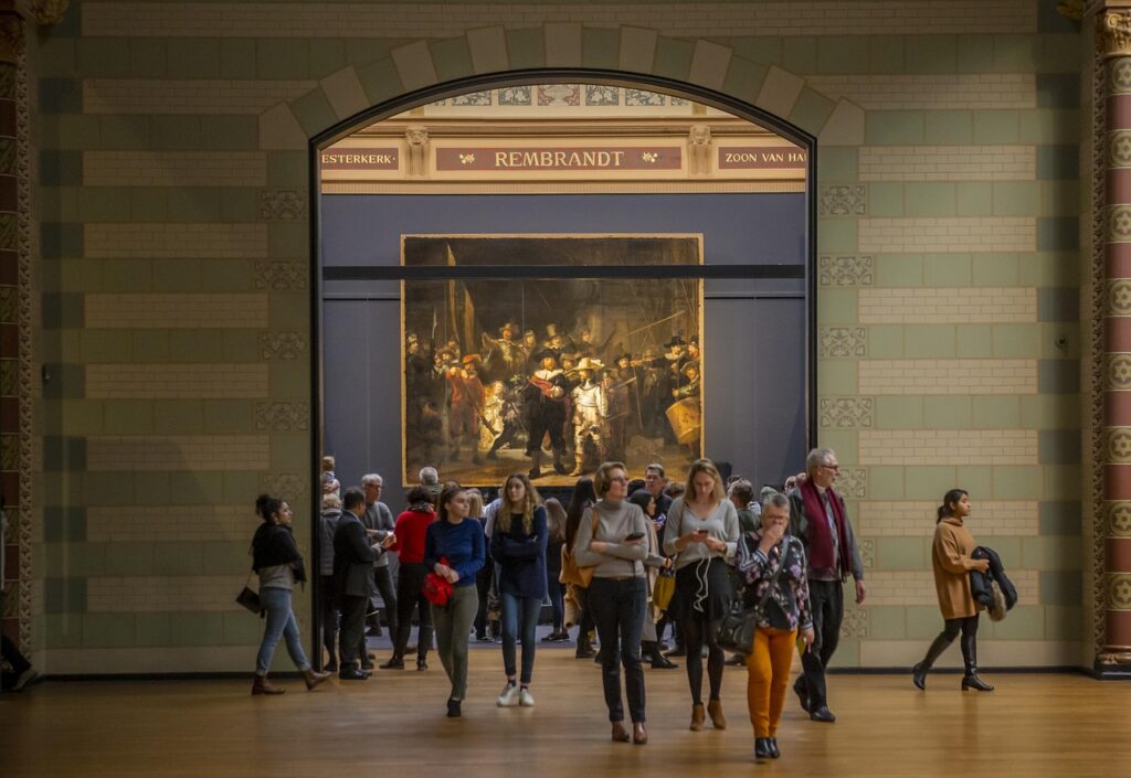 Een groep mensen, misschien op zoek naar hun ideale partner in de kunst, loopt en staat voor Rembrandts "De Nachtwacht", tentoongesteld in een grote, goed verlichte galerie met gedessineerde muren en een gewelfde deuropening.