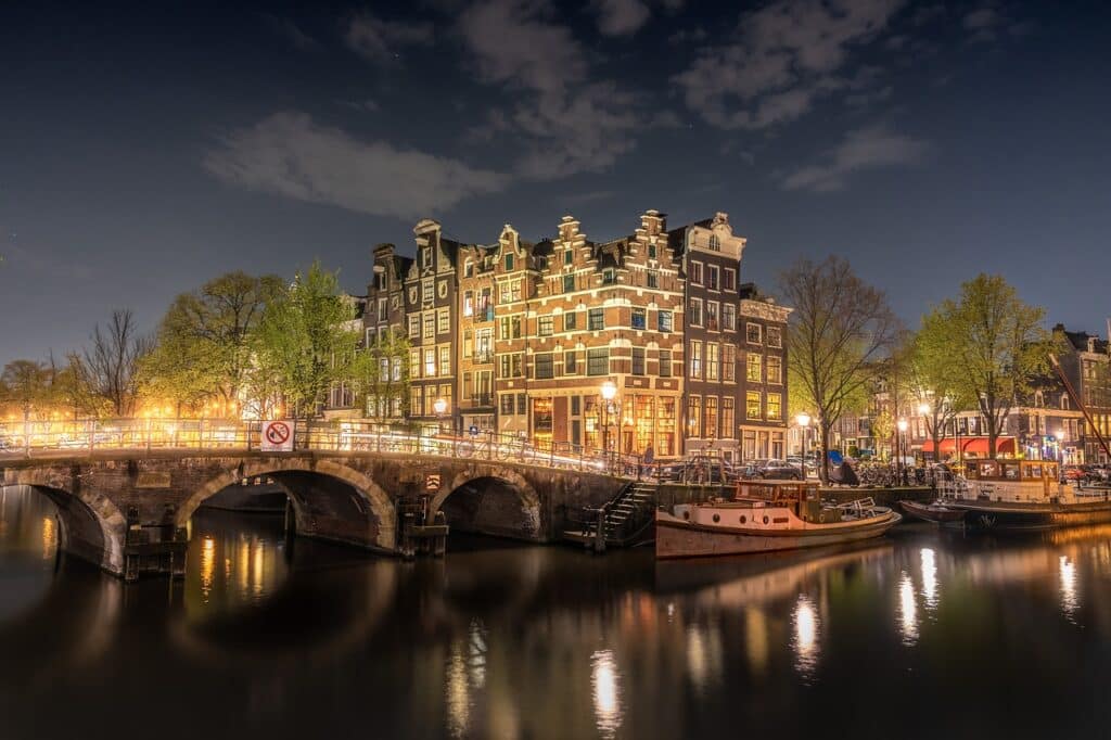 Nachtelijk uitzicht op de Amsterdamse gracht met een stenen brug, verlichte traditionele gebouwen met puntgevels en boten aangemeerd langs het water - een ideale partner voor een romantische wandeling onder bomen verlicht door straatverlichting onder een gedeeltelijk bewolkte hemel.