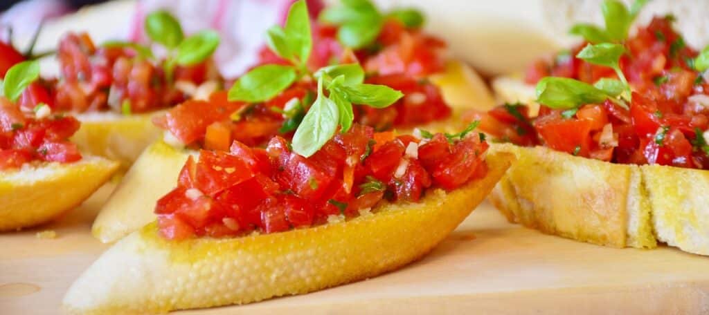 Close-up van verschillende bruschetta-plakken op een houten plank, belegd met blokjes tomaat, knoflook en verse basilicumblaadjes. Het brood ziet er knapperig en goudbruin uit, en is een perfect voorbeeld van hoe levendige rode en groene toppings een smakelijk contrast creëren.