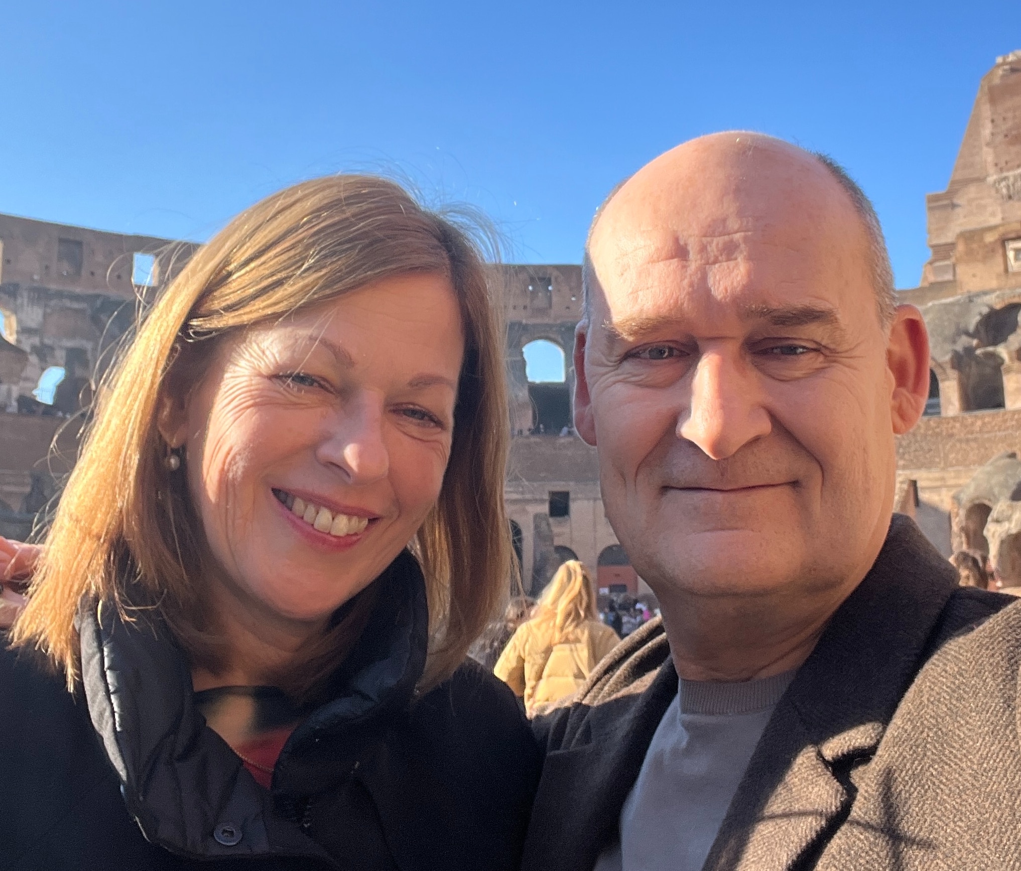 Patricia & Xavier 1 Een man en een vrouw met een hoog EQ omhelzen elkaar buiten, de ogen gesloten, terwijl de man zachtjes haar voorhoofd kust in een gebaar van troost en genegenheid. Beiden dragen blauw en zien er kalm en vredig uit tegen een achtergrond van groen.