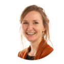 Een vrouw met lichtbruin haar, draagt een oranje-bruin jasje en een licht topje, glimlacht naar de camera tegen een effen witte achtergrond.