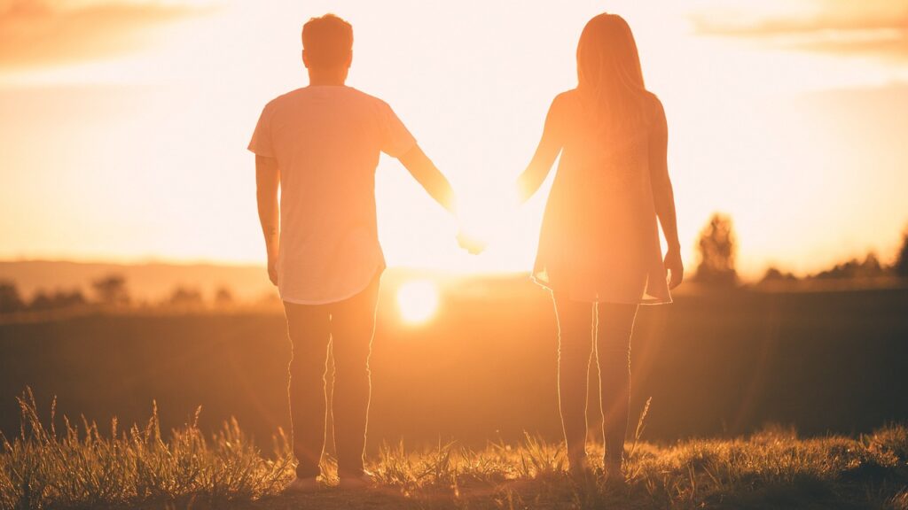 Een paar staat hand in hand, met uitzicht op een heldere zonsondergang in een open veld. Hun silhouetten gloeien in het warme licht en roepen hoop en genezing op na een relatie met een narcist, in een vredige en romantische sfeer.