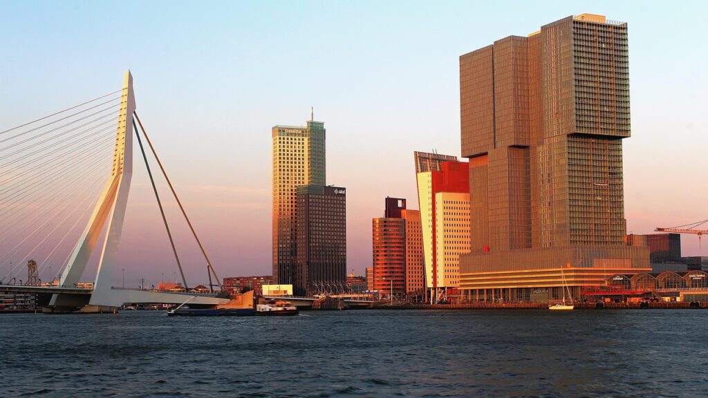 Een skyline van de stad bij zonsondergang met moderne hoogbouw, een tuibrug en boten op een rivier op de voorgrond - perfect voor date ideeën Rotterdam als het gouden licht het tafereel baadt in warmte en charme.