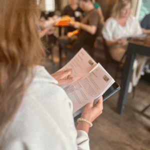 Een persoon leest een PartnerSelect-boekje in een lichte, informele omgeving terwijl anderen aan tafels op de achtergrond zitten en ervaringen uitwisselen of lezen. De focus ligt op het boekje en de handen van de persoon.