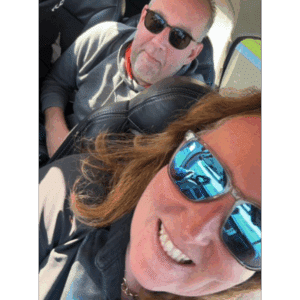 Een vrouw met een zonnebril lacht naar de camera terwijl ze een selfie neemt in een voertuig en dit moment van liefde deelt. Een man met een zonnebril zit achter haar en kijkt ook naar de camera terwijl het zonlicht weerkaatst op hun glazen - een ervaring om nooit te vergeten.