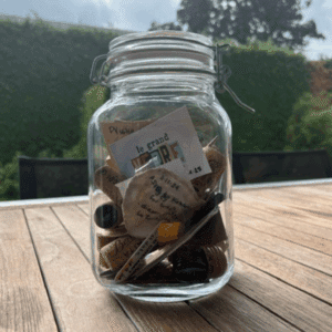 Een glazen pot gevuld met verschillende kleine voorwerpen, waaronder wijnkurken, briefjes en kaartjes - elk een herinnering aan liefde en ervaring - staat op een houten tafel buiten met groen en bomen op de achtergrond.