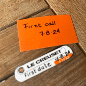 Een oranje kaart met "First call 7.8.24" erop geschreven ligt op een houten oppervlak naast een Le Creuset etiket met de tekst "first date 7.8.24" met een rood hart - perfecte herinneringen aan een ervaring vol liefde, geïnspireerd door PartnerSelect.