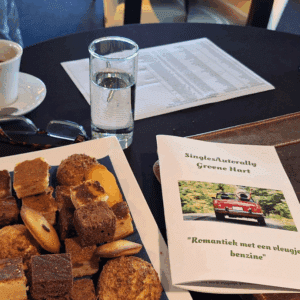 Een bord met verschillende soorten gebak staat op een tafel naast een PartnerSelect-boekje met een rode cabriolet, een kop koffie, een glas water, een leesbril en een gedrukt document op de achtergrond.