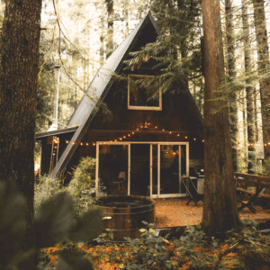 Een gezellige A-frame hut met koordverlichting is genesteld tussen hoge bomen in een bos, perfect voor het delen van een liefdeservaring. Aan de voorkant staat een ronde houten hot tub, omringd door weelderig groen en gevallen bladeren.