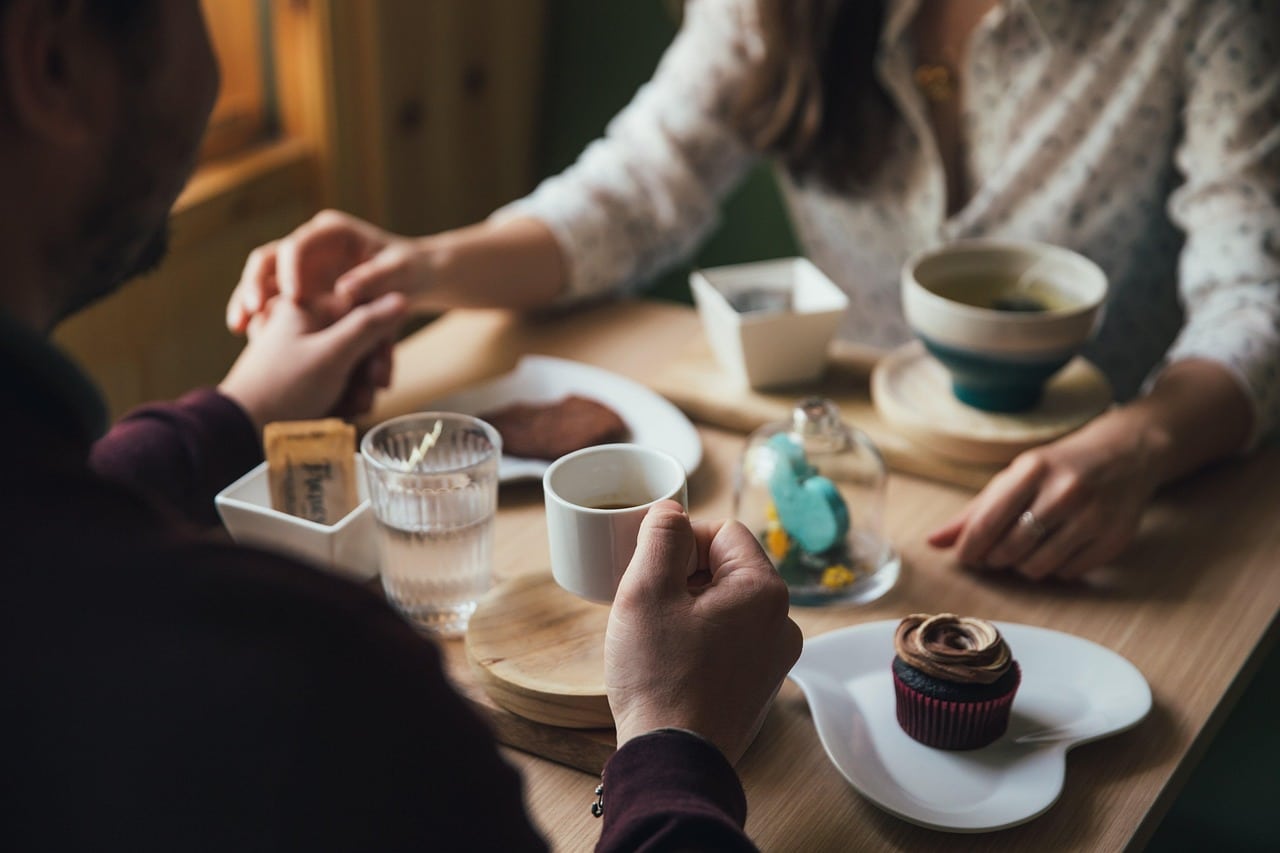 Een stel zit aan een houten tafel, houdt elkaars handen vast en deelt koffie, thee, een cupcake en gebak. De scène voelt warm en intiem aan en biedt inspiratie en datingtips voor wie op zoek is naar gezellige daten-momenten in natuurlijk licht. Een stel zit aan een houten tafel, houdt elkaars handen vast en deelt koffie, thee, een cupcake en gebak. De scène voelt warm en intiem aan en biedt inspiratie en datingtips voor wie op zoek is naar gezellige daten-momenten in natuurlijk licht.