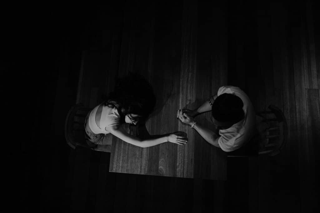 Een zwart-wit overheadfoto van twee mensen die tegenover elkaar zitten aan een houten tafel; de een steekt zijn hand uit, terwijl de ander met de handen in elkaar geklemd zit, de angst vastleggend om alleen te zijn na een relatie.