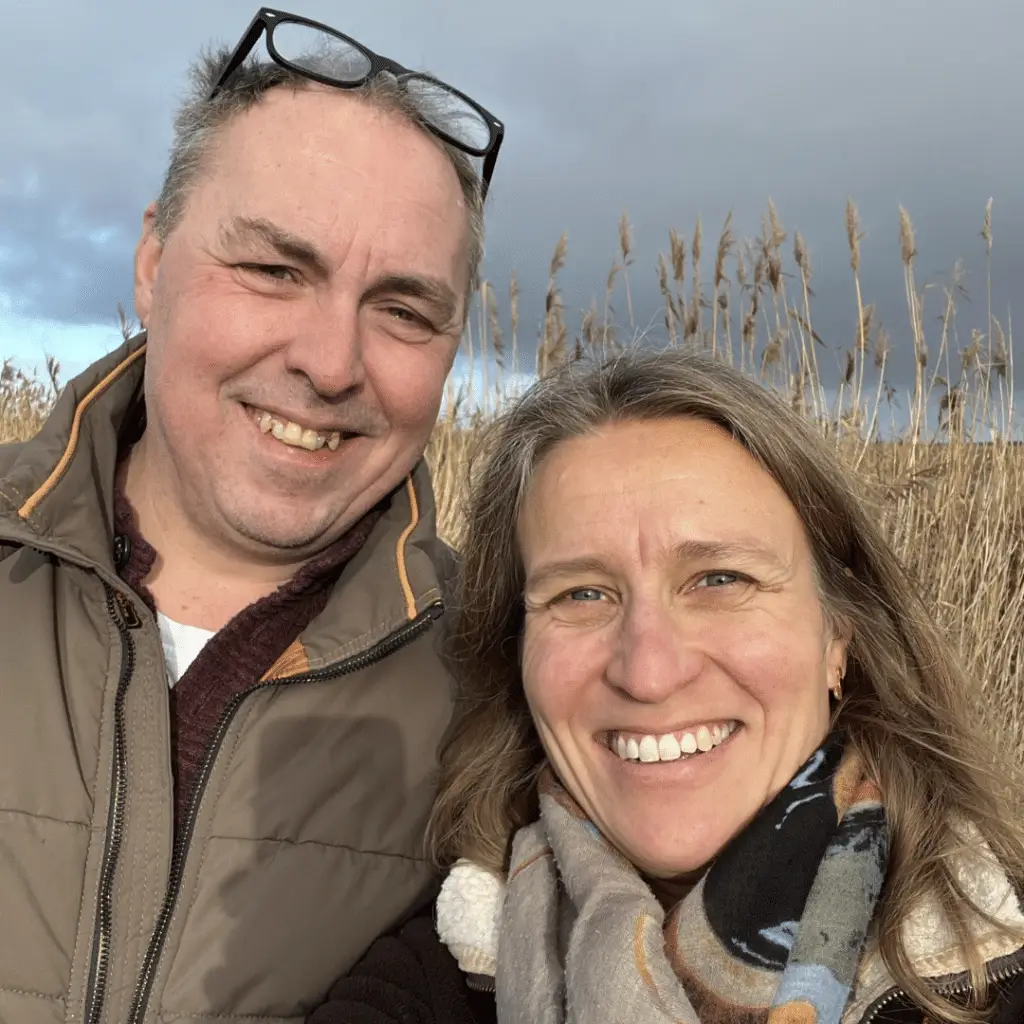 Dawn & Derek 1 Een man en een vrouw met een hoog EQ omhelzen elkaar buiten, de ogen gesloten, terwijl de man zachtjes haar voorhoofd kust in een gebaar van troost en genegenheid. Beiden dragen blauw en zien er kalm en vredig uit tegen een achtergrond van groen.