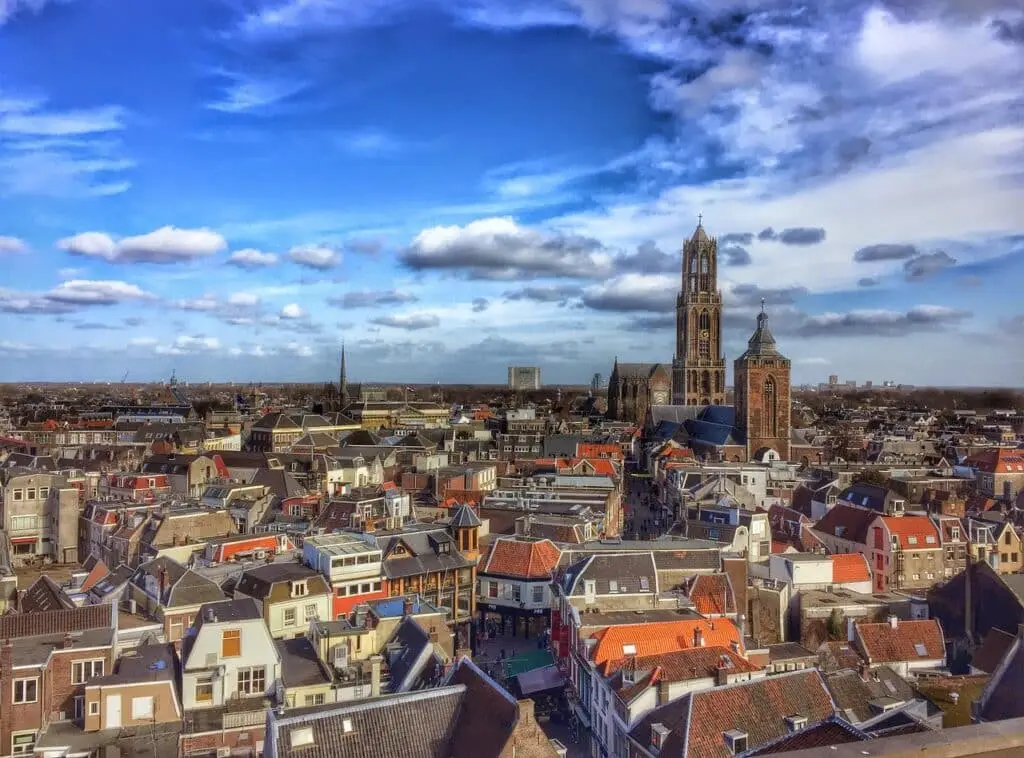 Romantiek in de Domstad: De leukste date ideeën in Utrecht 3 Een panoramisch uitzicht op Utrecht met dicht op elkaar gepakte huizen met rode en grijze daken, een hoge historische kerktoren en een blauwe lucht met verspreide wolken - perfect voor romantiek Utrecht of de leukste date ideeën Utrecht.