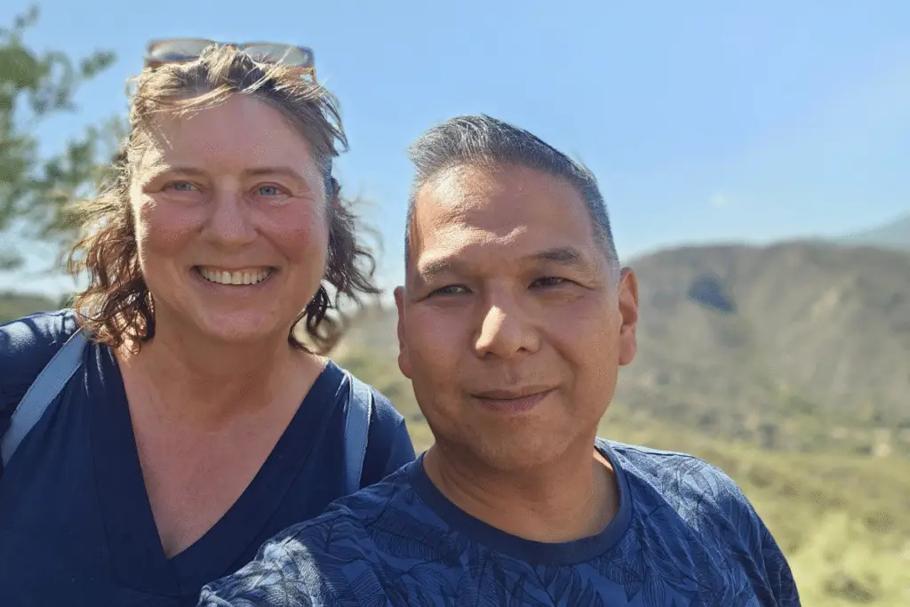 Jolanda & Koen 4 Twee mensen glimlachen buiten in het felle zonlicht en staan voor een schilderachtig, heuvelachtig landschap met blauwe lucht. Ze dragen allebei een marineblauw topje en delen hun ervaring met PartnerSelect. De persoon links heeft schouderlang haar en een bril op zijn hoofd.