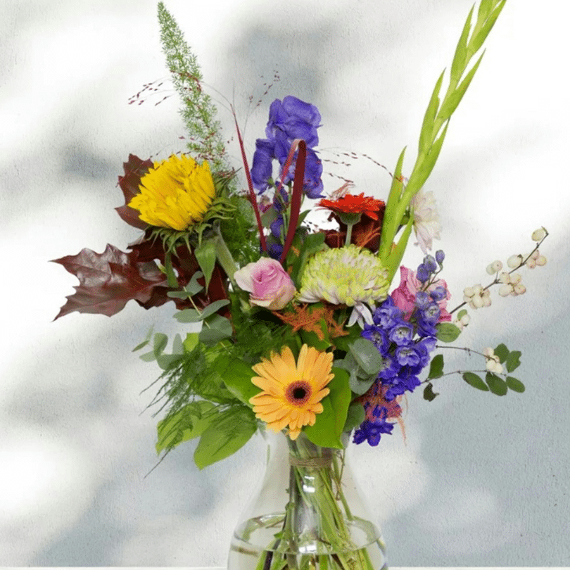 Een levendig boeket van diverse bloemen, waaronder zonnebloemen, rozen, gerbera margrieten en gladiolen, door PartnerSelect geschikt in een heldere glazen vaas tegen een lichte achtergrond met gevlekte schaduwen voor een onvergetelijke ervaring.