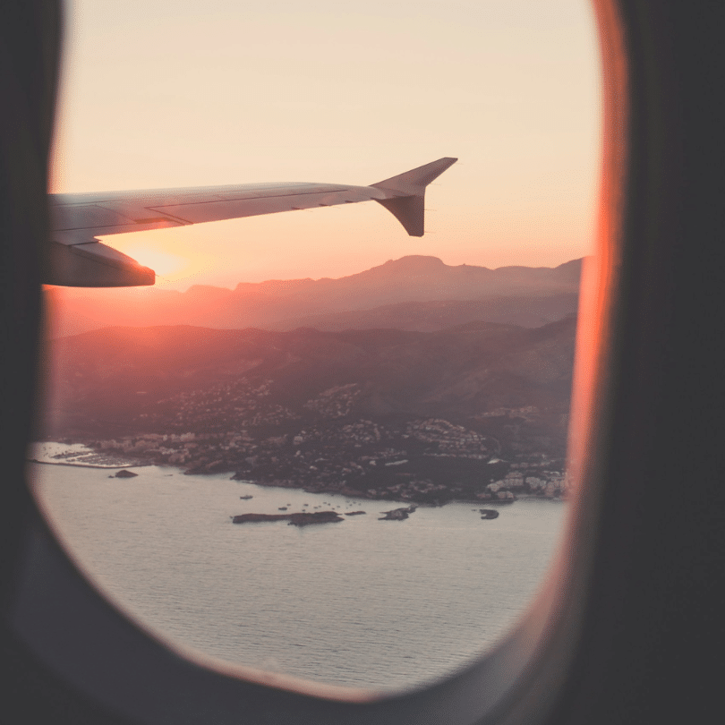 Uitzicht vanuit een vliegtuigraam op een vleugel, bergen en kustlijn bij zonsondergang, met warm, zacht licht dat een gloed over het landschap en het water werpt - een ervaring die doet denken aan de hoge reizen van PartnerSelect.