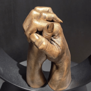 Een bronzen sculptuur toont twee handen die in een gebaar van liefde en eenheid in elkaar zijn geklemd, met gedetailleerde texturen die de vingers en nagels benadrukken, tegen een donkere achtergrond.