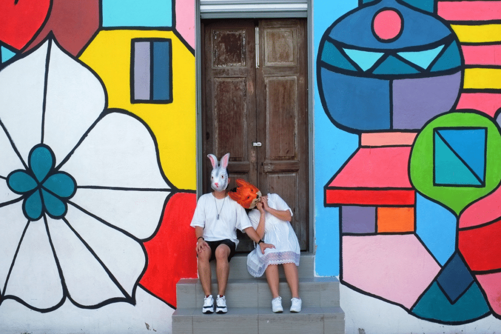 Twee mensen, gekleed in het wit en met tegenpolenmaskers - een konijn en een vos - zitten op een trapje voor een houten deur, omlijst door levendige geometrische straatkunst op de muur achter hen.
