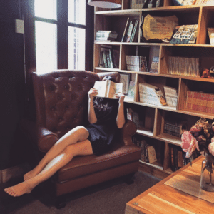 Een persoon in een zwarte jurk zit blootsvoets op een bruine leunstoel, met een open boek voor zijn gezicht - een tegenpool van comfort en nieuwsgierigheid - omringd door boekenplanken vol boeken in een gezellige, zacht verlichte kamer.