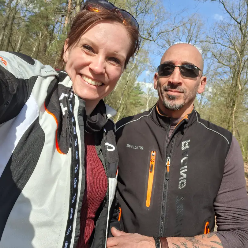 Marieke & Regev 1 Een man en een vrouw met een hoog EQ omhelzen elkaar buiten, de ogen gesloten, terwijl de man zachtjes haar voorhoofd kust in een gebaar van troost en genegenheid. Beiden dragen blauw en zien er kalm en vredig uit tegen een achtergrond van groen.