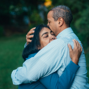Een man en een vrouw met een hoog EQ omhelzen elkaar buiten, de ogen gesloten, terwijl de man zachtjes haar voorhoofd kust in een gebaar van troost en genegenheid. Beiden dragen blauw en zien er kalm en vredig uit tegen een achtergrond van groen.