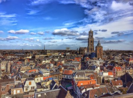 Een panoramisch uitzicht op Utrecht met dicht op elkaar gepakte huizen met rode en grijze daken, een hoge historische kerktoren en een blauwe lucht met verspreide wolken - perfect voor romantiek Utrecht of de leukste date ideeën Utrecht.