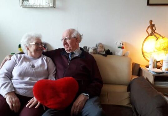 Een glimlachend ouder echtpaar zit op een bank, terwijl de vrouw een rood hartvormig kussen vasthoudt. Ze zijn omringd door knuffelbeesten en een warmgele lamp verlicht het tafereel vanaf de rechterkant. Dit vrolijke en tevreden stel, samengebracht door een vooraanstaande matchmaker, geniet van hun gezellige woonkamer.