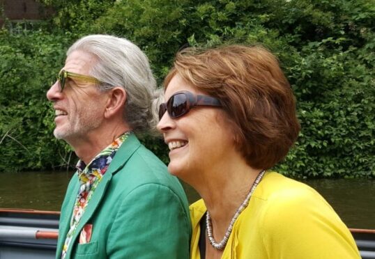 Twee oudere volwassenen, een man en een vrouw, zitten buiten aan een rivier, allebei glimlachend en met een zonnebril op. De man heeft grijs haar, een baard en draagt een groene blazer met een kleurrijk overhemd. De vrouw heeft kort bruin haar en draagt een geel topje met een ketting. Ze hebben elkaar ontmoet via een deskundige matchmaking-service.