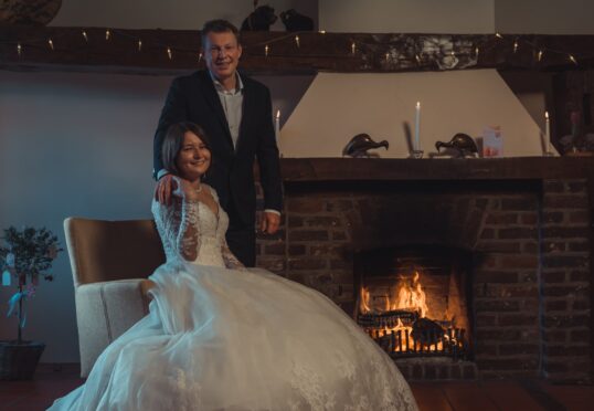 Een stel poseert in een elegante setting voor een relatiebemiddelingsadvertentie. De vrouw, gekleed in een witte trouwjurk, zit op een stoel met haar hand op de arm. De man, gekleed in pak, staat achter haar met zijn hand op haar schouder. Ze staan voor een verlichte bakstenen open haard.