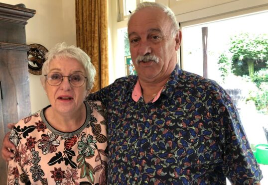 Een grijsharige vrouw met een bril en een gebloemde blouse staat naast een oudere man met een snor, gekleed in een overhemd met kleurrijke patronen. In de lichtgekleurde kamer van het matchmakingbureau stroomt zonlicht door een groot raam, waardoor groen op de achtergrond zichtbaar wordt.