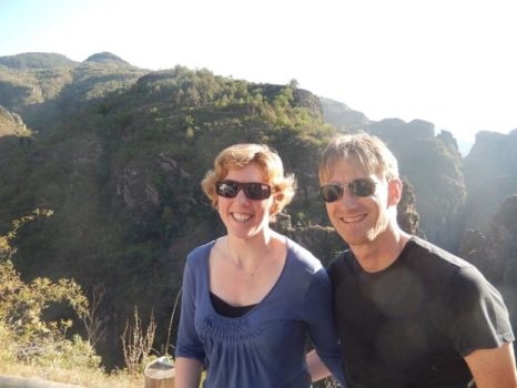 Een glimlachende vrouw en man, beiden met een zonnebril, staan buiten dicht bij elkaar tegen een achtergrond van zonovergoten bergen en groen. De vrouw draagt een blauw topje en de man een zwart overhemd. De zon schijnt fel op de achtergrond, wat duidt op het begin van hun duurzame relatie, gevoed door deskundige relatiebemiddeling.