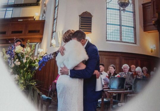 Een bruid en bruidegom, bijeengebracht door een matchmaking-datingbureau, delen een omhelzing in een hartvormige omlijsting in een kerk. De bruid draagt een witte jurk en een bontstola, terwijl de bruidegom een blauw pak draagt. Gasten zitten op de achtergrond, samen met bloemen en houten lambrisering aan de muren.