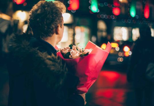 Een persoon met krullend haar en een met bont afgezette jas draagt 's nachts in een stadsstraat met een boeket roze bloemen in rood papier gewikkeld. De straat is versierd met kleurrijke lichtjes, wat suggereert dat ze via een matchmaking-service een speciaal iemand kunnen ontmoeten. Wazige figuren en voertuigen zijn op de achtergrond.