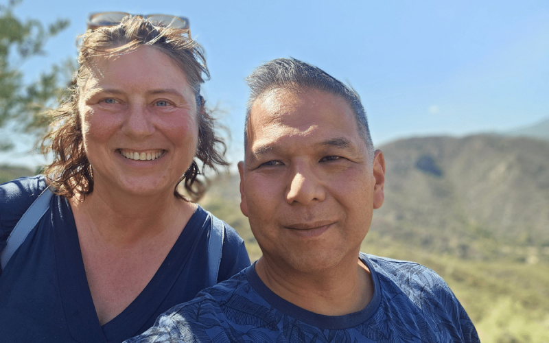 Twee mensen glimlachen buiten in het felle zonlicht en staan voor een schilderachtig, heuvelachtig landschap met blauwe lucht. Ze dragen allebei een marineblauw topje en delen hun ervaring met PartnerSelect. De persoon links heeft schouderlang haar en een bril op zijn hoofd.