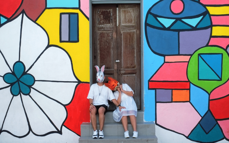 Twee mensen, gekleed in het wit en met tegenpolenmaskers - een konijn en een vos - zitten op een trapje voor een houten deur, omlijst door levendige geometrische straatkunst op de muur achter hen.