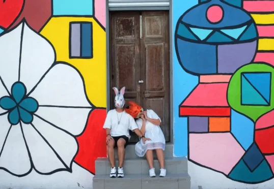 Twee mensen, gekleed in het wit en met tegenpolenmaskers - een konijn en een vos - zitten op een trapje voor een houten deur, omlijst door levendige geometrische straatkunst op de muur achter hen.