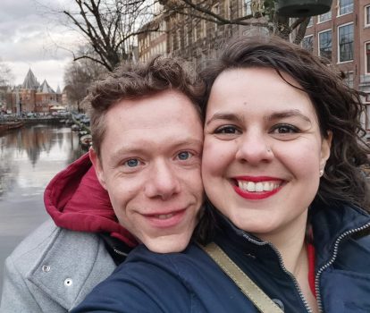 Een glimlachend koppel maakt een close-up selfie langs een gracht in een stad met historische gebouwen op de achtergrond. De man heeft licht haar en draagt een grijze jas met een rode capuchon, terwijl de vrouw donker haar, rode lippenstift heeft en marineblauw gekleed is. Hun vrolijke uitdrukking doet vermoeden dat ze elkaar via een datingbureau hebben ontmoet.
