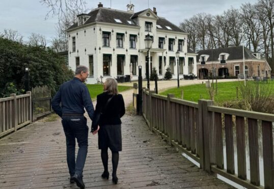 Een stel loopt hand in hand over een houten brug naar een groot, wit huis van twee verdiepingen met groene luiken. Op de achtergrond zijn kale bomen en bewolkte luchten te zien. Het huis is omgeven door een groen gazon en nog een kleiner gebouw, wat de charme oproept die vaak wordt geassocieerd met traditionele relatiebemiddeling.